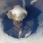 A volcanic eruption on an island sending a thick grey ash cloud into the sky above the coast.