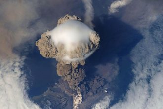 A volcanic eruption on an island sending a thick grey ash cloud into the sky above the coast.